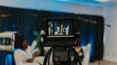 A video camera records a woman talking at a live event on stage.
