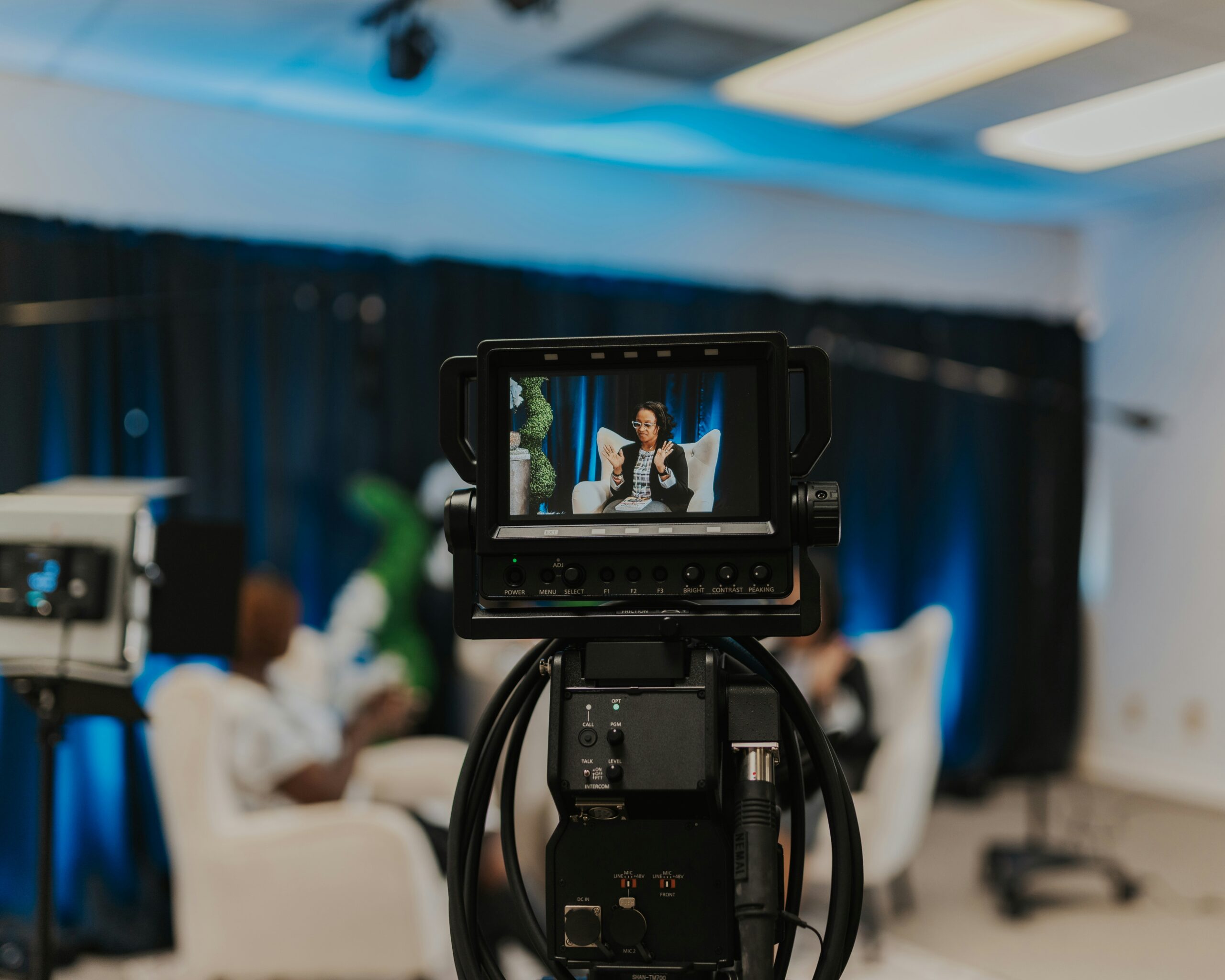 A video camera records a woman talking at a live event on stage.